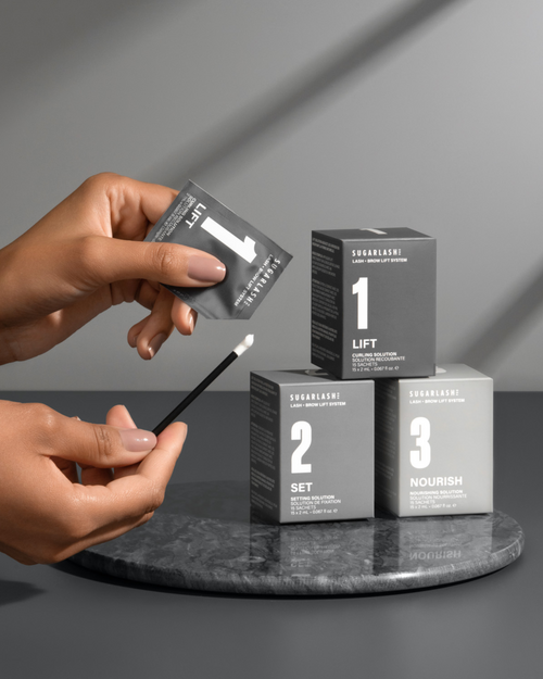 Model holding a Lift sachet near Lift, Set, and Nourish Lash and Eyebrow Lamination boxes.