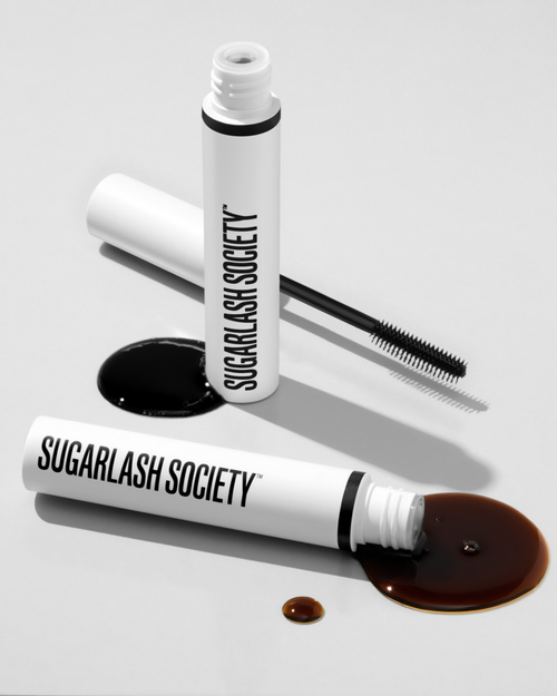 Brown and black glaze tubes with product for lash extensions spilled out on white background.