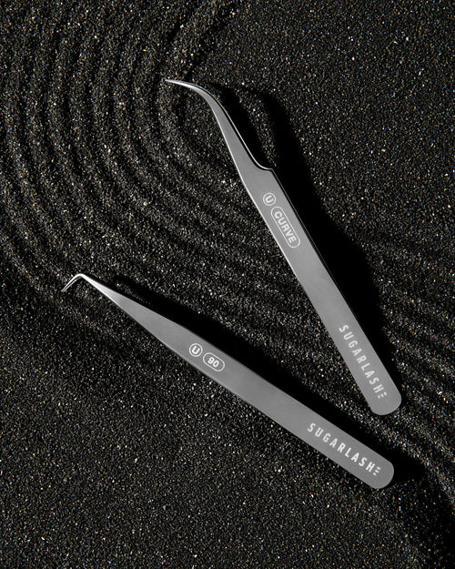 Two lash extension Tweezers laid on black and metallic sand.