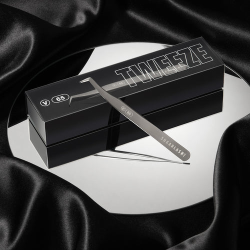 A box of lash extensions Volume lash Tweezer V-65 on top of a mirror with an unboxed pair of tweezers resting partially on the box.