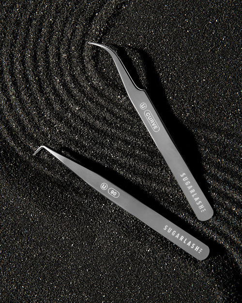 Two Lash extension Tweezers laid on black and metallic sand.